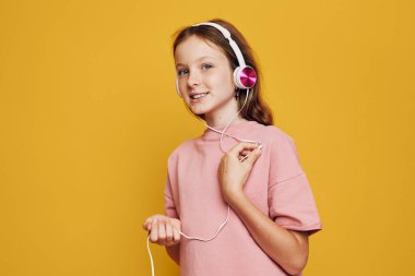 Pembe kulaklıklı, açık pembe tişörtlü, elinde kulaklık tutarken gülümseyen kızıl saçlı kız. Parlak sarı arka plan neşeli, modern atmosfer yaratır. Müzik ve gençlik kavramı.