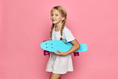 Turkuaz kaykay taşıyan mutlu sarışın kız pembe bir duvarın önünde duruyor. O, hafif bir günlük kıyafet giydi. Açık hava aktiviteleri, gençlik ve oyun kavramı.