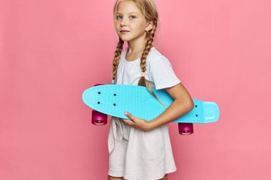 Saçları örgülü sarışın kız pembe bir arka fonun önünde mavi bir kaykayla poz veriyor. O, beyaz bir gömlek ve bej şort giydi. Gençlik, eğlence ve spor anlayışı.