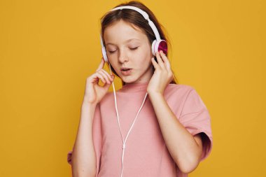 Kulaklıklı pembe tişörtlü genç kız müzikten hoşlanıyor. Gözleri kapalı, sarı arka planda odaklanmış bir ifade. Müzik, gençlik ve rahatlama konseptleri için stüdyo çekimi.
