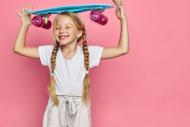 Saçları örgülü, mavi kaykayı başının üstünde tutan sevimli, gülümseyen kız pembe arka planda duruyor. Çocukluk mutluluğu kavramı. Eğlence ve dışarıda zaman geçirme kavramı..