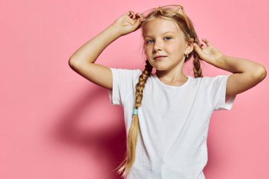 Beyaz tişörtlü, örgülü ve gözlüklü kendine güvenen genç kız pembe bir duvarın önünde duruyor. Parlak stüdyo ışıkları onun doğal ifadesini ve duruşunu vurguluyor..