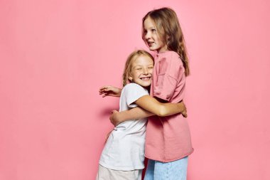 Yüzlerinde gülücüklerle kucaklaşan iki neşeli kızın portresi. Pastel pembe bir arka planda. Stüdyo aydınlatmasında arkadaşlık, şefkat ve kardeş bağı kavramı.