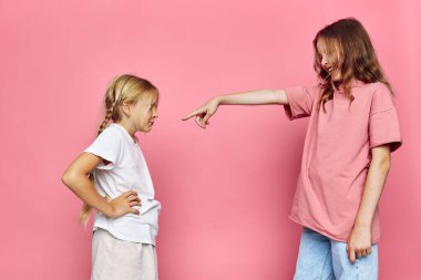 İki çocuk pembe bir zemine karşı duruyor. Kızlardan biri saldırganca işaret ediyor, diğeri rahatsız edici bir şekilde tepki veriyor ve kollarını kavuşturuyor. Çatışma, gerginlik ve anlaşmazlık kavramı.