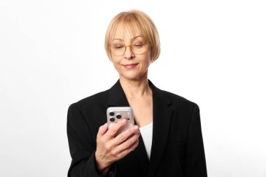 Gözlüklü ve siyah ceketli kendinden emin olgun bir kadın beyaz arka planda hafif bir gülümsemeyle akıllı telefona bakar. Teknoloji kullanımı, iletişim, modern yaşam tarzı kavramı.