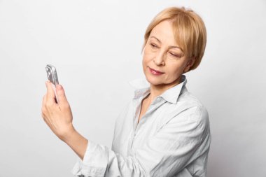 Sıradan, orta yaşlı, kısa sarı saçlı bir kadın beyaz arka planda özçekim yapmak için akıllı telefon kullanıyor. Teknoloji kavramı, dijital iletişim ve günlük yaşam.