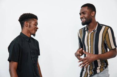 Siyah tenli iki siyah adam ayakta ve gülümserken beyaz arka planda günlük kıyafetlerle arkadaşça sohbet ediyorlar. Genç yetişkinler arasında sosyal bağlantı ve mutluluk.