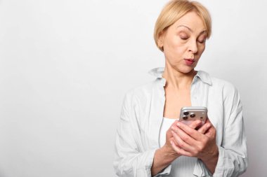 Kısa saçlı, beyaz gömlekli sarışın kadın akıllı telefon tutuyor ve şaşırmış görünüyor. Beyaz arka planda izole edilmiş, teknoloji, iletişim, duygu ve günlük yaşam tarzı kavramı..