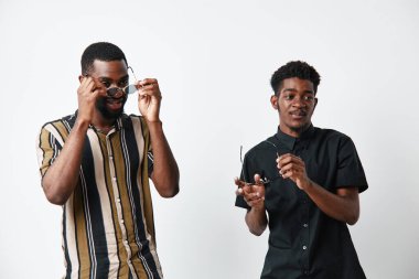 Siyah tenli, güneş gözlüğü tutan iki Afro-Amerikalı adamın portresi. Beyaz arka planlı stüdyo çekimi, gündelik kıyafetler ve modern tarzda rahat bir kendine güven havası..