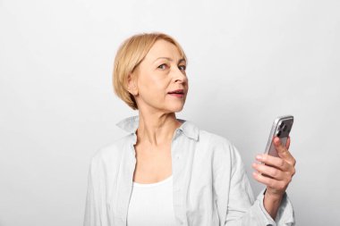 Kendine güvenen yetişkin bir kadının portresi. Kısa sarı saçlı, akıllı bir telefon tutan ve yana bakan. Beyaz gömlek ve sade arkaplan, gündelik tarz ve modern teknoloji kullanımını vurguluyor.