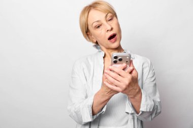 Beyaz gömlek giyen orta yaşlı bir kadın akıllı telefon tutuyor ve ağzı açık bir şekilde şaşkınlık gösteriyor. Beyaz arka planda izole edilmiş. Şok, iletişim, tepki ve teknoloji kavramı.
