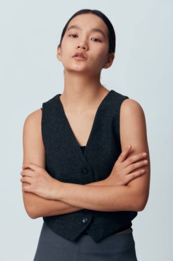 Kolları çapraz duran genç bir kadın kolsuz siyah yelek giyiyor. Nötr yüz ifadesi, temiz arka plan, sıradan zarif tarz, kapalı mekan fotoğrafı, doğal makyaj, pürüzsüz cilt.