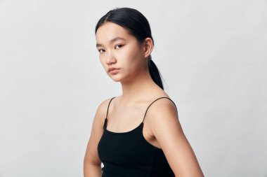 Siyah kolsuz bluzlu, saçları arkadan bağlanmış ciddi bir genç kadının portresi. Minimalist stüdyo ayarları tarafsız zemin üzerinde yüz hatlarını ve gündelik modern kıyafetleri vurgular.