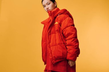 Kırmızı kış kirpi ceketli ve turuncu arka planda poz veren katmanlı bir kadın model. Şık dış giyim, rahat kıyafet, havalı tavır ve renkli minimalist stüdyo portresi..