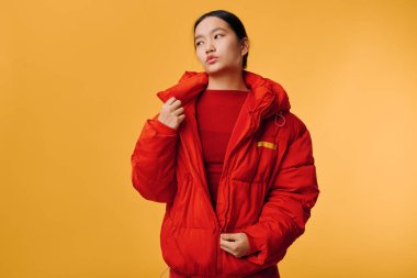 Parlak kırmızı ceketli genç kadın sarı arka planda poz veriyor. Soğuk mevsim için sıcak giysili moda portresi ve modern fotoğraf stüdyosunda düşünceli bir ruh hali..