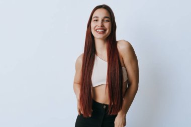 Uzun kahverengi saçlı, beyaz kolsuz bluzlu ve siyah kotlu gülümseyen bir kız. Doğal görünüşü ve rahat bir tarzı olan mutlu bir kadının portresi..