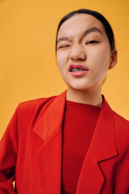 Kırmızı kıyafetli bir kadın model. Bir gözü açık, parlak sarı arka planda şakacı bir şekilde göz kırpıyor. Kendine güvenen yüz ifadesini ve modern günlük kıyafet tarzını gösteren yakın plan portre..