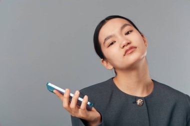 Akıllı telefonu tutan ve arka planda kameraya bakan kendine güvenen genç bir kadının portresi. Stüdyo ortamında odaklanmış ve sakin giyinen kadın model..