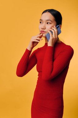 Uzun siyah saçlı, kırmızı kurdeleli bir elbise giyen, sarı stüdyo geçmişi, iletişim ve cihaz kullanımı için cep telefonuyla konuşan düşünceli genç bir kadının portresi..