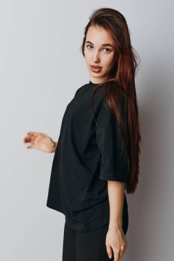 Uzun düz saçlı, siyah tişörtlü ve pantolonlu, stüdyo ortamında gri arka planda kameraya bakan genç kadın modelin portresi..