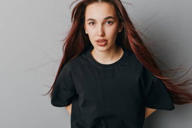 Siyah tişörtlü, dalgalı kahverengi saçlı genç bir kadının portresi. Gri arka planda poz veriyor. Minimalist stüdyo ortamında doğrudan bakıyor ve sakin bir yüz ifadesi sergiliyor..