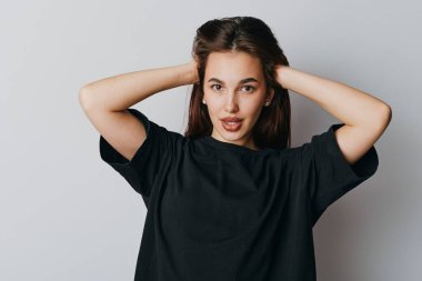 Siyah gömlekli, kolları başının arkasında poz veren genç bir kadın kameraya kendinden emin bir yüz ifadesi ve tarafsız gri arka planda doğal ışık ile bakıyor..