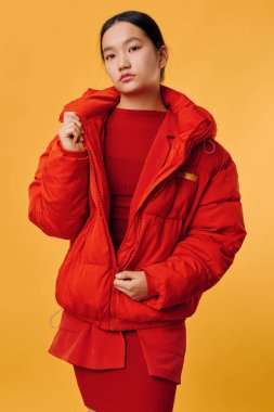 Parlak kırmızı kabarık ceket giyen ve aynı elbiseyi giyen şık bir kadın fotoğraf stüdyosundaki gündelik kıyafetlerle sarı zemine karşı güvenle poz veriyor..