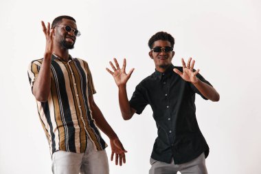 Siyah tenli, şık giyinmiş, güneş gözlüklü, beyaz arka planda dans eden ve gülümseyen Afrikalı etnik erkekler. Genç arkadaşlar içeride neşe ve pozitif enerji ifade ediyorlar..