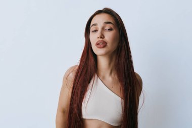 Tarafsız arkaplan önünde duran uzun saçlı ve beyaz bluzlu genç bir kadın portresi, sakin bir yüz ve doğal güzelliği ev ortamında minimum makyajla gösteriyor..