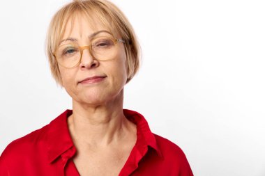Gözlüklü, kırmızı bluzlu, özgüvenli orta yaşlı bir kadının beyaz arka planda kameraya baktığı yakın plan. Yaşlanma, kendine güven, bilgelik ve doğal güzellik kavramı.