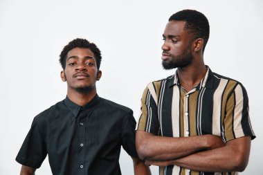 İki koyu tenli Afro-Amerikan erkek portresi, biri siyah gömlekli, diğeri de kolları çapraz çizgili çizgili, beyaz arka planda ciddi ifadelerle poz veriyor..
