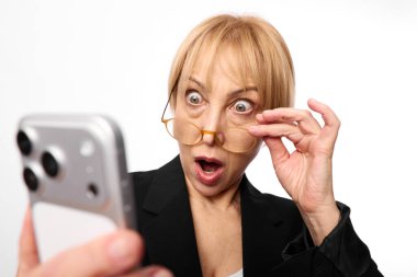 Siyah ceketli, şok olmuş orta yaşlı bir kadın, beyaz arka planda izole bir şekilde akıllı telefona bakarken gözlüklerini indiriyor. Şaşırtma, güvensizlik ve teknoloji kavramı.