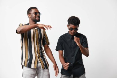 Siyah tenli, güneş gözlüklü Afro-Amerikalı erkekler dans etmekten zevk alıyorlar ve beyaz stüdyo arka planında çizgili ve siyah tişörtleri var..