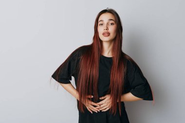 Siyah tişört giyen, uzun düz saçlı genç bir kadının portresi. Kadın, elleri kalçasında kameraya bakıyor. Beyaz arka planda tarafsız bir yüz var..