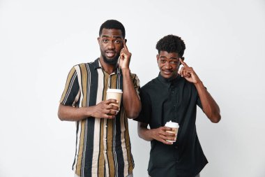 Koyu tenli Afrikalı erkekler kahve fincanları ve dokunaklı kafalarla ayakta duruyorlar. Arkadaşlık, günlük kıyafetler ve beyaz stüdyo arka planında düşünceli bir ifade..