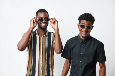 Siyah tenli, siyah saçlı, güneş gözlüğü takan ve günlük gömlekler giyen iki siyah adamın portresi. Yaşam tarzı ve gençlik kültürü için beyaz arka planda duran gülümseyen arkadaşlar.