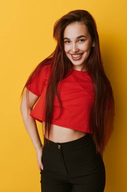 Kırmızı gömlekli, siyah pantolonlu, sarı arka planda poz veren kendinden emin genç bir kadın. Uzun saçlı, gülümseyen, cebinde fotoğraf çekimi için duran bir manken..
