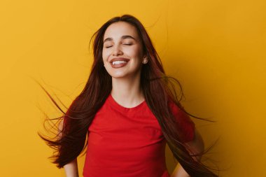 Parlak sarı duvarın önünde gülümseyen dalgalı saçları ve kapalı gözleri olan mutlu kız. Kırmızı tişört giyen, neşe ve sükuneti ifade eden, tasasız tavırlı ve yeni görünüşlü bir kadın..