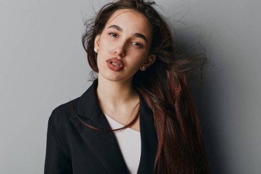 Uzun saçlı, siyah ceketli ve beyaz gömlekli genç bir kadının portresi. Stüdyo aydınlatma ortamında gri duvarın yanında poz veriyor..