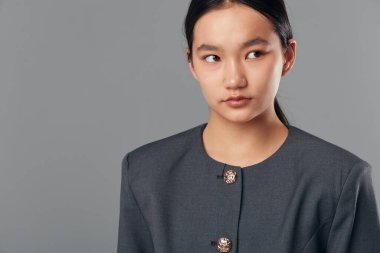 Siyah saçlı, gri resmi ceketli, yana bakan genç bir Asyalı kadının portresi. Pürüzsüz ten, düzgün saç, minimum makyaj, basit arka plan, iş ya da moda kavramı.