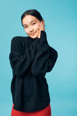 Mavi duvarın önünde eli nazikçe birbirine dokunan siyah kazaklı ve kırmızı etekli kadın model. Doğal makyajlı genç Asyalı kadın günlük moda tarzı için bol örgü giyiyor.