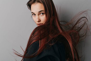 Dalgalı kahverengi saçlı ve siyah elbiseli genç bir kadının portresi. Gri arka planda ciddi bir yüze sahip kameraya bakıyor. Güzellik, moda ve saç hareketleri stüdyoda doğal olarak çekildi.