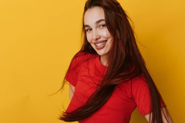 Kıvırcık saçlı, kırmızı gömlekli, canlı sarı arka planda duran gülümseyen genç bir kadın. Neşeli ifade ve rahat görünüm gösteren kadın modeli..