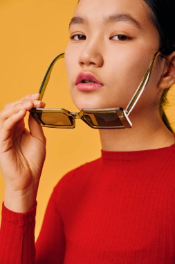 Kırmızı bluz giyen, sarı arka planda güneş gözlüğü tutan genç bir kadın. Modern tarz ve kendine güvenen yüzüyle moda çekimi için doğal makyajlı sıradan görünümlü bir güzellik portresi..