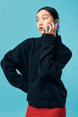 Siyah örgü süveter ve kırmızı etek giyen bir kadın stüdyoda konuşmak için kulağına yakın bir yerde akıllı telefon tutuyor. Dijital aygıt ve mavi arkaplan ile portre, iletişim ve modern yaşam tarzı.