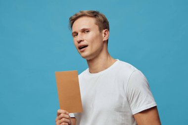 Gündelik beyaz tişörtlü beyaz genç adam kahverengi kâğıt nota basıyor. Mavi arka planda düşünceli bir ifadeyle yan yana bak. Dijital göçebe yaşam tarzı kavramı, akıllı telefon kullanımı, uzaktan çalışma ve