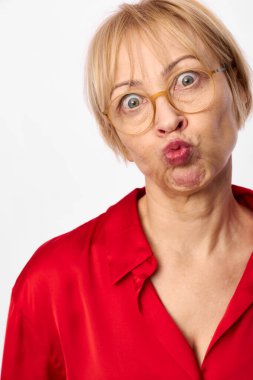 Sarışın, orta yaşlı, gözlüklü ve kırmızı gömlekli bir kadın dudaklarını büzüyor. Beyaz arkaplanlı yakın plan portre, neşeli ruh hali, mizah ve yaşam tarzı konsepti için neşeli duygular..