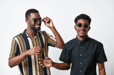 Siyah tenli ve güneş gözlüklü, gülümseyen ve eğlenen iki Afrikalı adamın portresi. Günlük kıyafet, pozitif ruh hali ve beyaz fon üzerine dostluk konsepti.