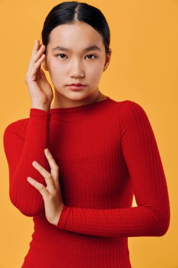 Siyah saçlı, kırmızı kazaklı, düşünceli bir bakışla kameraya bakan bir manken. Parlak turuncu arkaplan, temiz tasarım ve zarif moda veya yaşam tarzı fotoğrafı için yüzün yanına yerleştirilmiş eller.