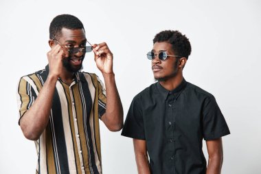 Siyah tenli, şık giyimli ve beyaz arka planda yan yana duran güneş gözlüklü iki Afrikalı adamın portresi..